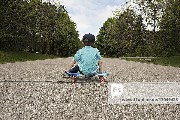 Rückansicht eines Jungen  der auf einem Skateboard am Fussweg sitzt
