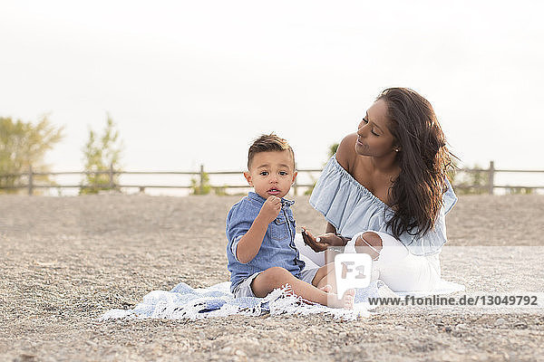 Porträt eines süßen Sohnes  der mit der Mutter auf einer Picknickdecke vor dem Himmel am Strand sitzt