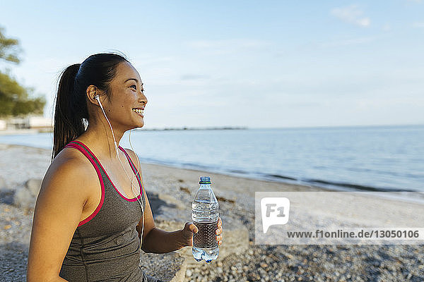 Happy woman listening music while holding water bottle at beach against sky