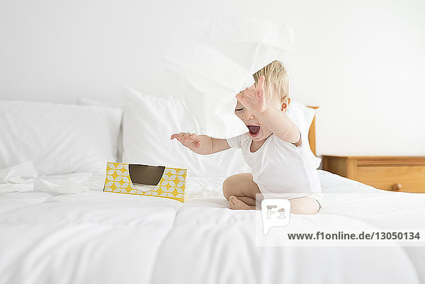 Fröhlicher Junge spielt zu Hause am Bett sitzend mit Gesichtstüchern
