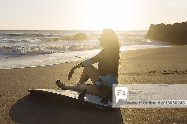 Seitenansicht einer Frau mit Surfbrett  die bei Sonnenuntergang am Strand vor klarem Himmel sitzt