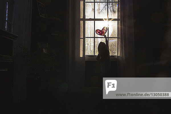Rear view of girl making heart shape on window at home