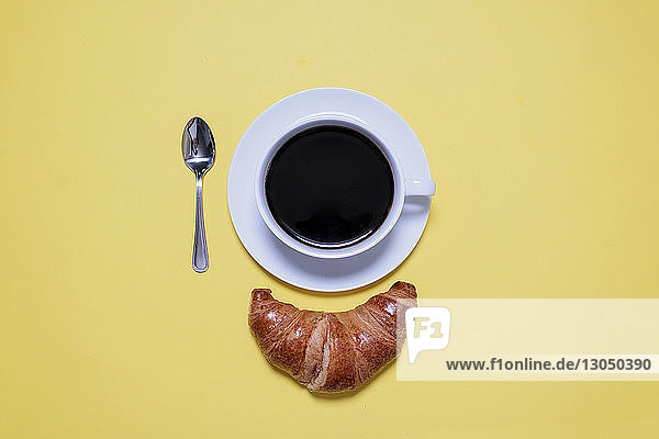 Draufsicht auf schwarzen Kaffee  Löffel und Croissant auf gelbem Tisch