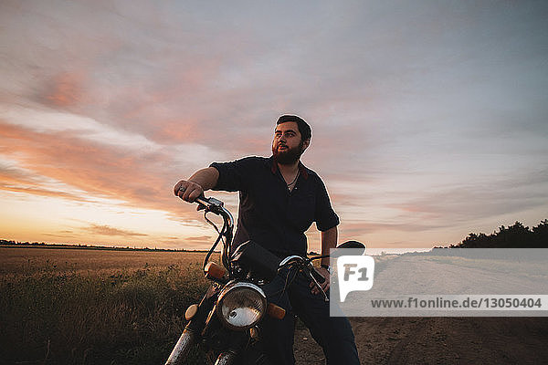 Nachdenklicher junger Mann sitzt auf einem Motorrad auf einem Bauernhof gegen den Himmel bei Sonnenuntergang