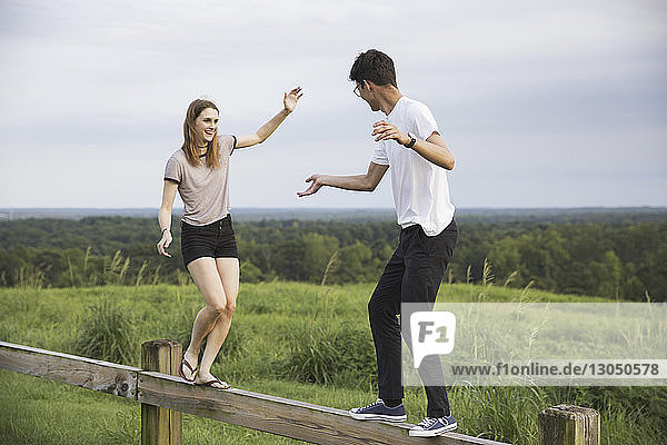 Happy friends walking on wooden railing over green landscape against sky