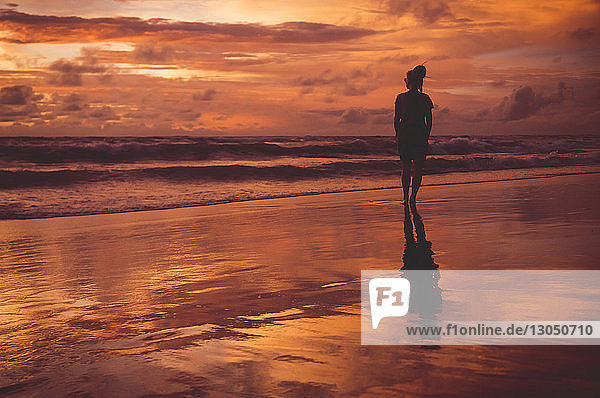 Rückansicht einer am Strand stehenden Frau vor dramatischem Himmel bei Sonnenuntergang