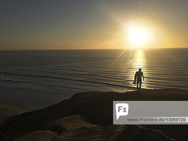 Silhouette man with surfboard standing on cliff by sea during sunset