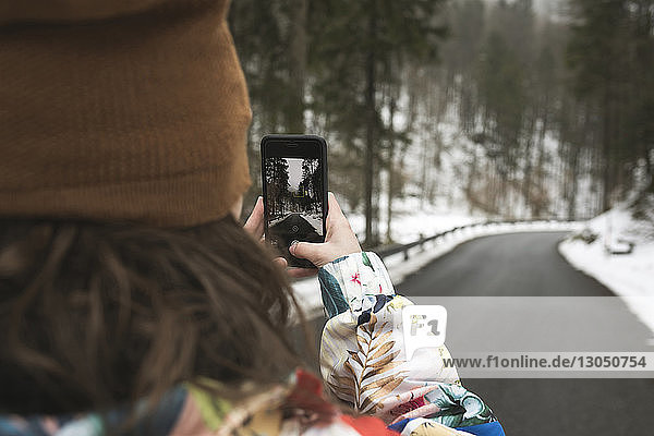 Rückansicht einer Frau  die im Winter im Wald eine Landstraße mit einem Smartphone fotografiert