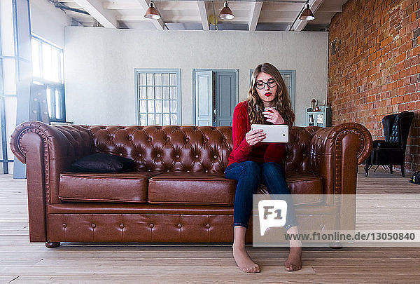 Full length of young woman using tablet computer while sitting on leather sofa at home