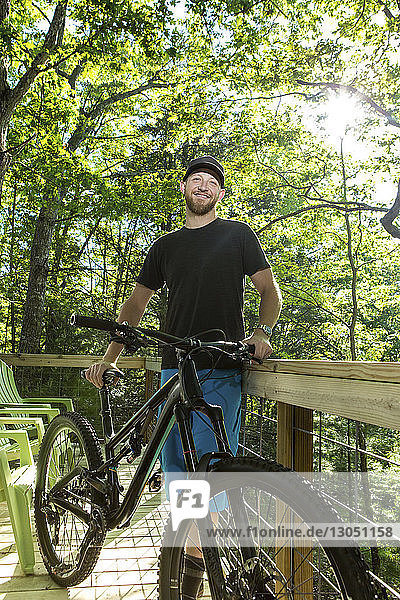 Portrait of confident hiker holding mountain bike while standing by fence at campsite in forest