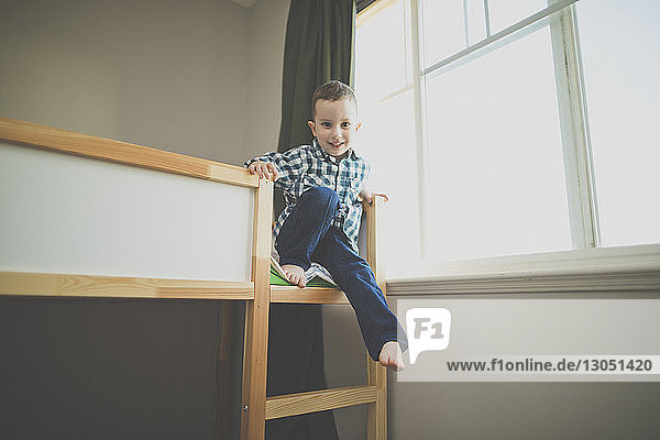Full length portrait of smiling boy sitting on bunk bed by window at home