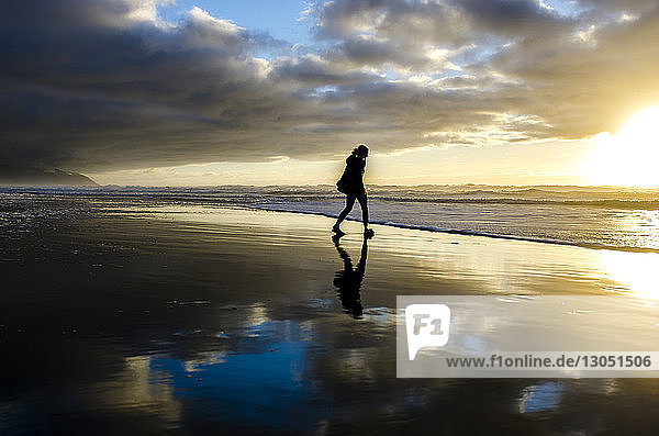 Silhouette einer Frau  die bei Sonnenuntergang am Strand vor bewölktem Himmel spazieren geht