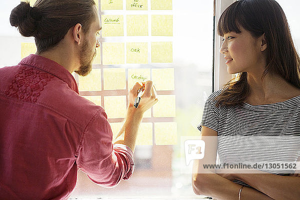 Businesswoman looking at colleague writing on adhesive note in creative office