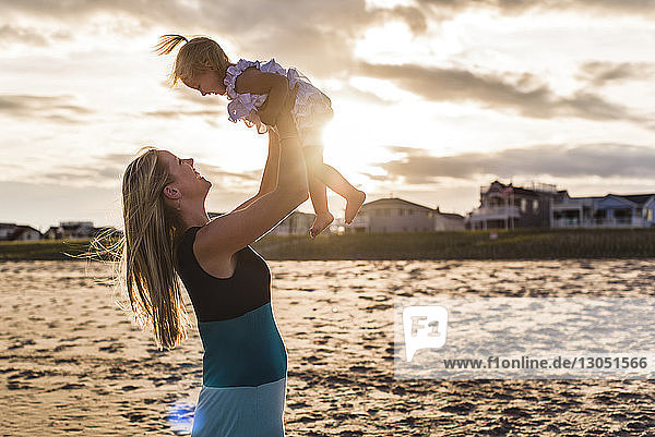 Seitenansicht einer spielerischen Mutter  die ihre Tochter beim Spielen am See gegen den bewölkten Himmel hebt