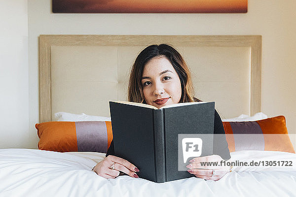 Portrait of confident businesswoman reading book on bed in hotel room