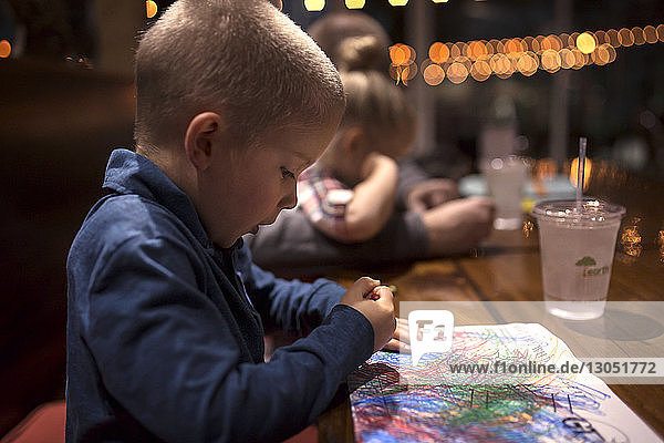 Side view of boy coloring on paper at dining table in restaurant