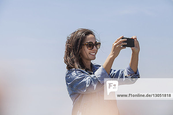 Niedrigwinkelansicht einer glücklichen Frau  die durch ein Smartphone vor klarem Himmel fotografiert
