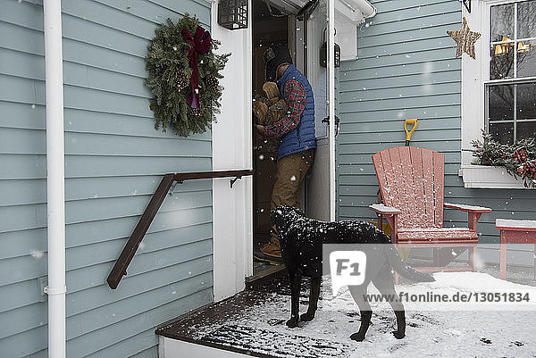 Mann mit Brennholz betritt das Haus  während der Hund an der Tür steht