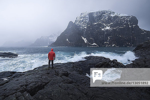 Rear view of man standing at rocky shore during foggy weather