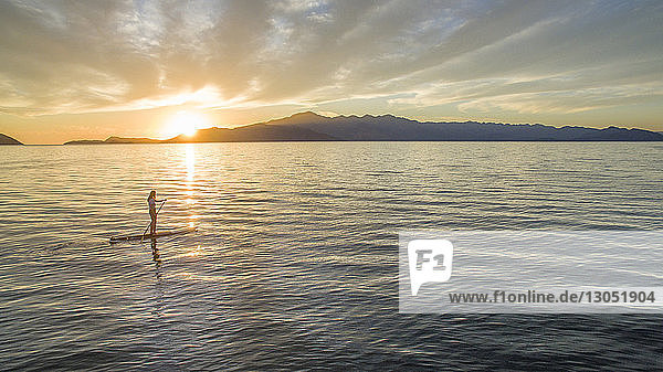 Frau paddelt im Meer gegen bewölkten Himmel bei Sonnenuntergang