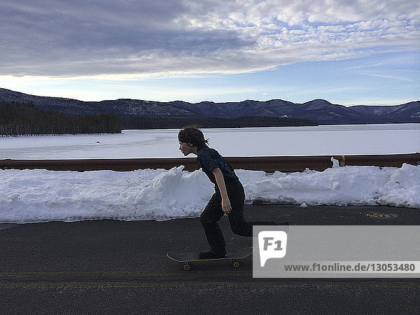 Seitenansicht in voller Länge eines Jungen,  der im Winter auf einer Straße am Fluss Skateboard fährt