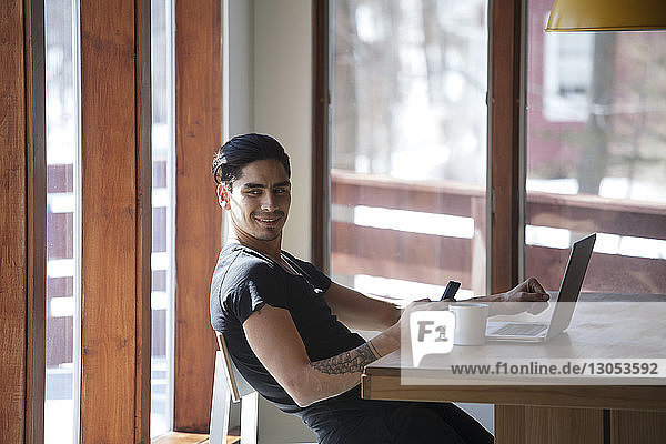 Lächelnder Mann mit Laptop-Computer  während er zu Hause auf einem Stuhl am Tisch sitzt