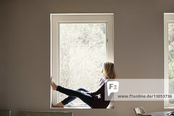 Seitenansicht einer nachdenklichen Frau  die zu Hause auf dem Fensterbrett sitzt