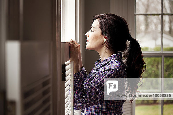 Side view of woman looking through window while standing at home