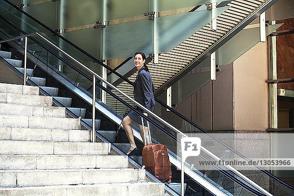 Selbstbewusste Geschäftsfrau steht mit Koffer auf der Rolltreppe