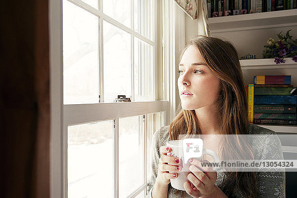 Nachdenkliche Frau hält Kaffeetasse  während sie zu Hause durchs Fenster schaut