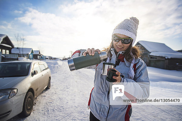 Glückliche Frau gießt Getränk aus Isolierkanne aus  während sie auf schneebedecktem Feld steht