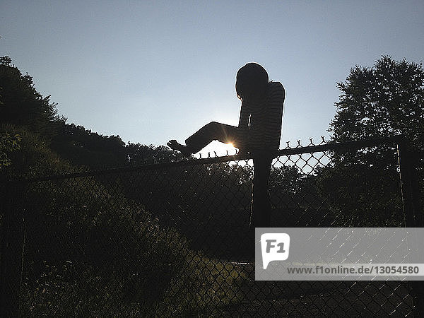 Junge klettert in voller Länge über den Kettengliedzaun im Feld gegen den klaren blauen Himmel