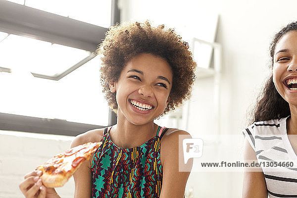Happy girl looking away while holding pizza while sitting by sister at home