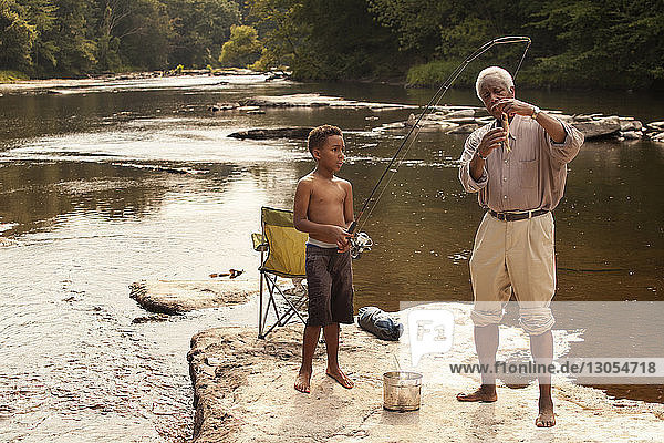 Boy holding fishing rod by grandfather looking at fish at lake