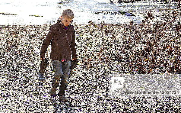 Boy carrying rubber boot while walking on field by river