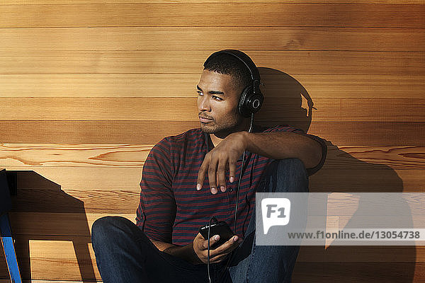 Man looking away while listening music on headphone against wall