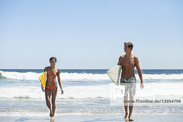 Glückliches Paar trägt Surfbrett beim Strandspaziergang vor klarem Himmel
