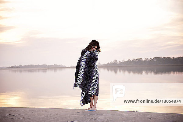 Woman wrapped in blanket while standing on beach against sky