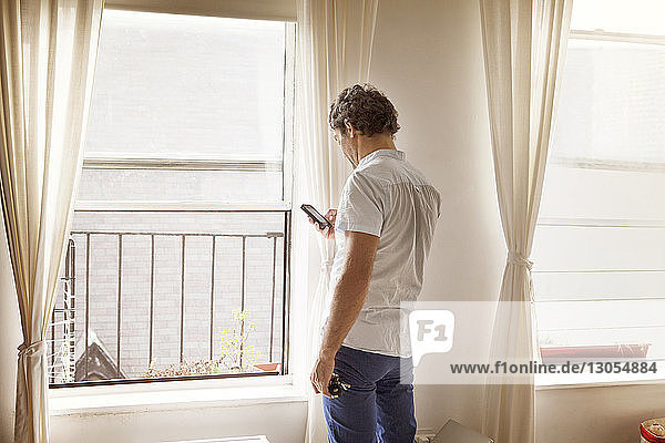 Mann benutzt Mobiltelefon  während er zu Hause am Fenster steht