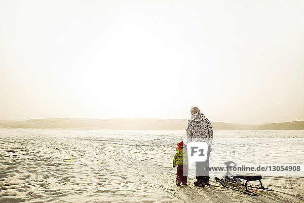 Rückansicht von Mutter und Tochter im Schlitten stehend auf schneebedecktem Feld vor klarem Himmel