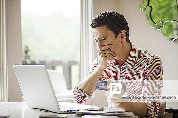 Besorgter Mann hält Kreditkarte  während er auf Laptop-Computer schaut