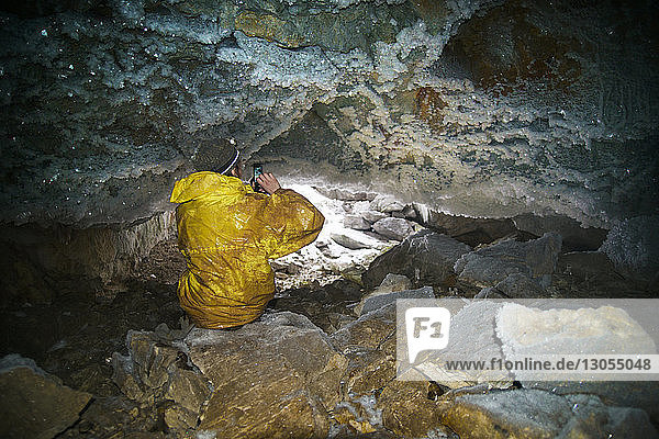 Rückansicht eines Mannes  der mit einem Mobiltelefon fotografiert  während er auf einem Felsen in einer Höhle sitzt