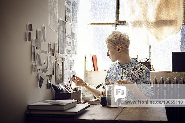 Woman sticking photographs on wall while sitting by table at home