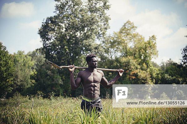 Shirtless muscular farmer with shovel standing on field during sunny day