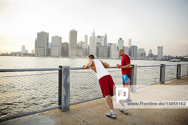 Father talking with man exercising on promenade by East River in city against sky