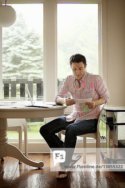 Mann liest Dokument  während er zu Hause auf einem Stuhl am Tisch sitzt