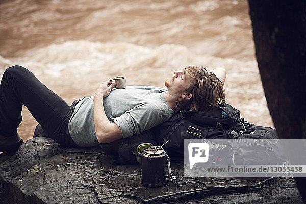 Man with mug sleeping on rock at riverbank