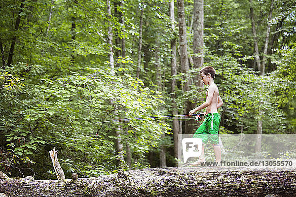 Seitenansicht eines Jungen mit Angelrute  der im Wald auf einem umgefallenen Baumstamm geht