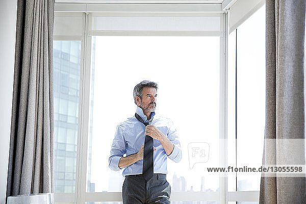 Businessman tying necktie while standing against window at hotel room