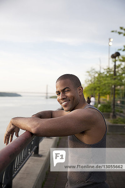 Portrait of happy man standing at railing in park by river against sky